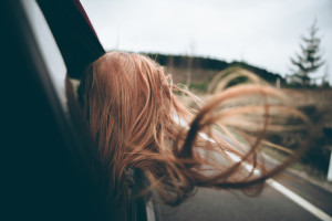 Frau streckt den Kopf aus dem Autofenster, ihre langen Haare fliegen im Fahrtwind